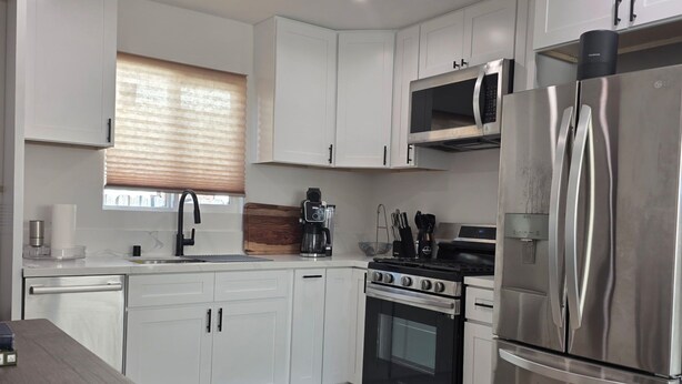 Una cocina moderna con armarios blancos, un refrigerador de acero inoxidable, un microondas, una estufa y una ventana con persianas.