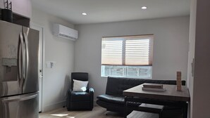 Una cocina moderna con refrigerador, un comedor con mesa y sillas, un sofá negro y una ventana con persianas.