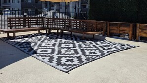 Un área de estar al aire libre con bancos de madera y sombrillas grandes, un tapete estampado y un seto al fondo.