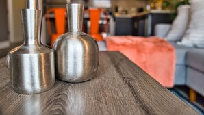 Twee metalen vazen op een houten tafel in een woonkamer, met een bank en een plaid op de achtergrond.