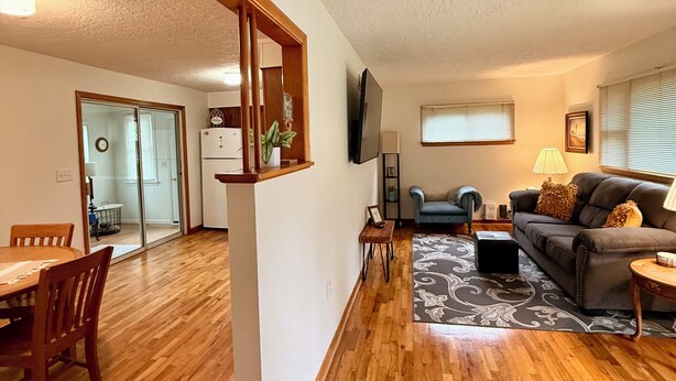 A spacious living room with hardwood flooring, a grey sofa, a coffee table, and a TV mounted on the wall. - Cozy 2-bedroom house in tranquil Excelsior Springs with AC (Excelsior Springs)