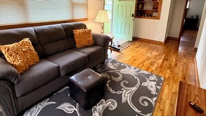 A living room with a sofa, ottoman, patterned rug, wooden floor, and a door with a 'hello' sign. - Cozy 2-bedroom house in tranquil Excelsior Springs with AC (Excelsior Springs)