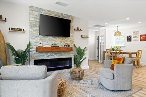 Ein Wohnzimmer mit Kamin aus Stein, einem Flachbildfernseher, grauen Sesseln und einem Essbereich mit Holztisch und Stühlen.