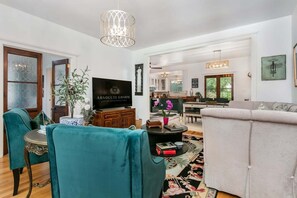 A spacious living room with teal chairs, a grey sofa, a flat-screen TV, and a chandelier. - Hackberry Haus | Hot Tub, Fire-pit, Pet Friendly w/ fee (Fredericksburg)