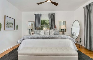 A bedroom with a bed, bedside tables, a ceiling fan, a window with curtains, and a mirror. - Hackberry Haus | Hot Tub, Fire-pit, Pet Friendly w/ fee (Fredericksburg)