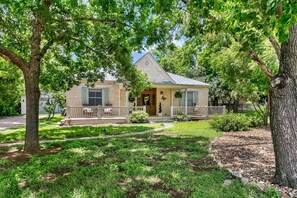 A two-story stone house with a porch, surrounded by lush greenery and trees. - Hackberry Haus | Hot Tub, Fire-pit, Pet Friendly w/ fee (Fredericksburg)