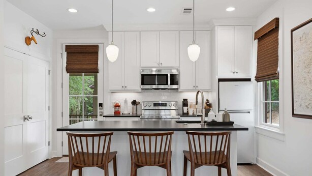 Una cocina moderna con armarios blancos, electrodomésticos de acero inoxidable, una isla central con taburetes y luminarias colgantes.