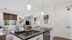 Una cocina moderna con encimera negra, muebles blancos y lavavajillas integrado. Se observan cuadros en la pared, una ventana con persianas y una puerta con picaporte decorativo.