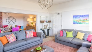 Ein Wohnzimmer mit zwei grauen Sofas, bunten Kissen, einem Chandelier, einem Essbereich mit Tisch und Stühlen sowie einem Gemälde an der Wand.