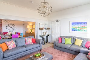 Ein Wohnzimmer mit zwei grauen Sofas, bunten Kissen, einem Chandelier, einem Essbereich mit Tisch und Stühlen sowie einem Gemälde an der Wand.