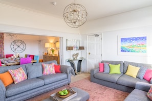 A living room with two grey sofas, colorful cushions, a chandelier, a dining area with a table and chairs, and a painting on the wall. - Northcliffe (8), Southwold, Suffolk (Southwold)