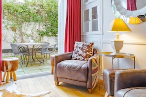 A room with a leather armchair, a small round table with a lamp, a dining table with chairs, and a view of a garden through a glass door. - Northcliffe (8), Southwold, Suffolk (Southwold)