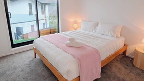 Een slaapkamer met een grote schuifpui, een bed met wit beddengoed, een roze plaid en een bedlampje.