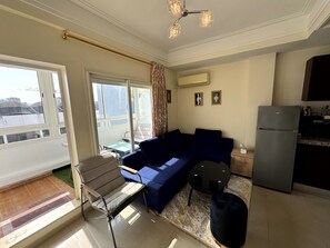 Ein Wohnzimmer mit blauem Sofa, einem kleinen Tisch, einem Stuhl, einem schwarzen Hocker, einer Küchenzeile mit Kühlschrank und einem Fenster mit Vorhängen.