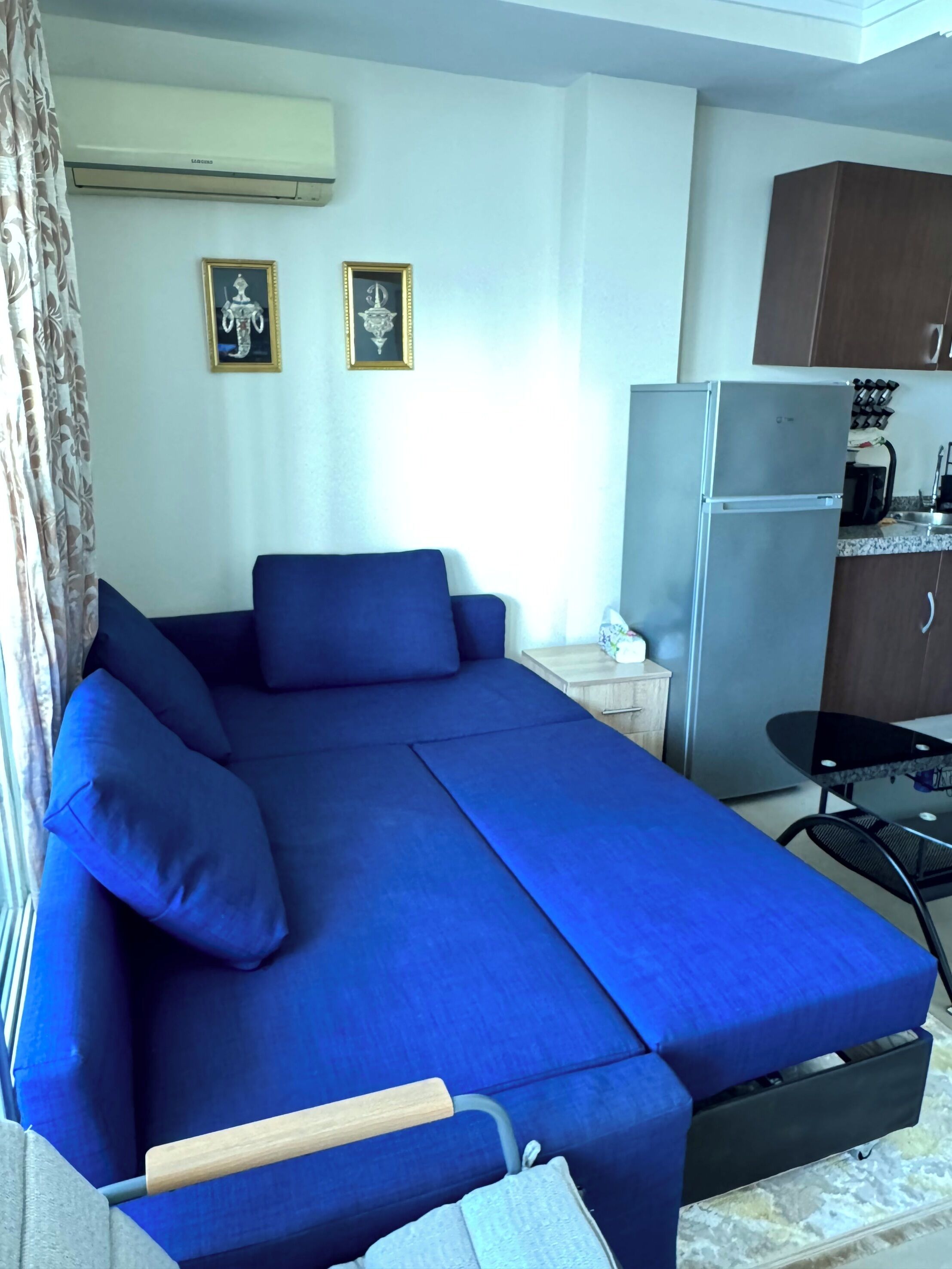 A compact room with a blue sofa, a small table, a refrigerator, and a window with curtains.
