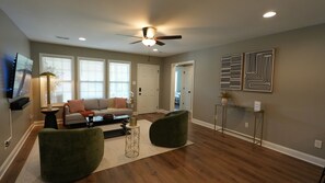 A living room with a sofa, coffee table, and chairs, a TV mounted on the wall, and a ceiling fan.