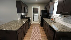 A kitchen with granite countertops, dark cabinets, a microwave, stove, dishwasher, and a checkered rug.