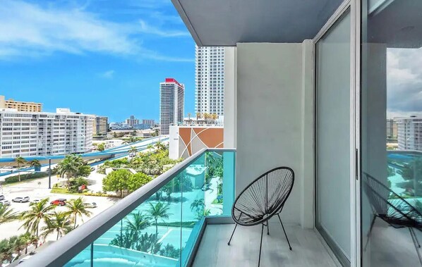 Ein Balkon mit Glasgeländer, einem Stuhl und Blick auf eine Stadt mit Gebäuden und einer Straße.