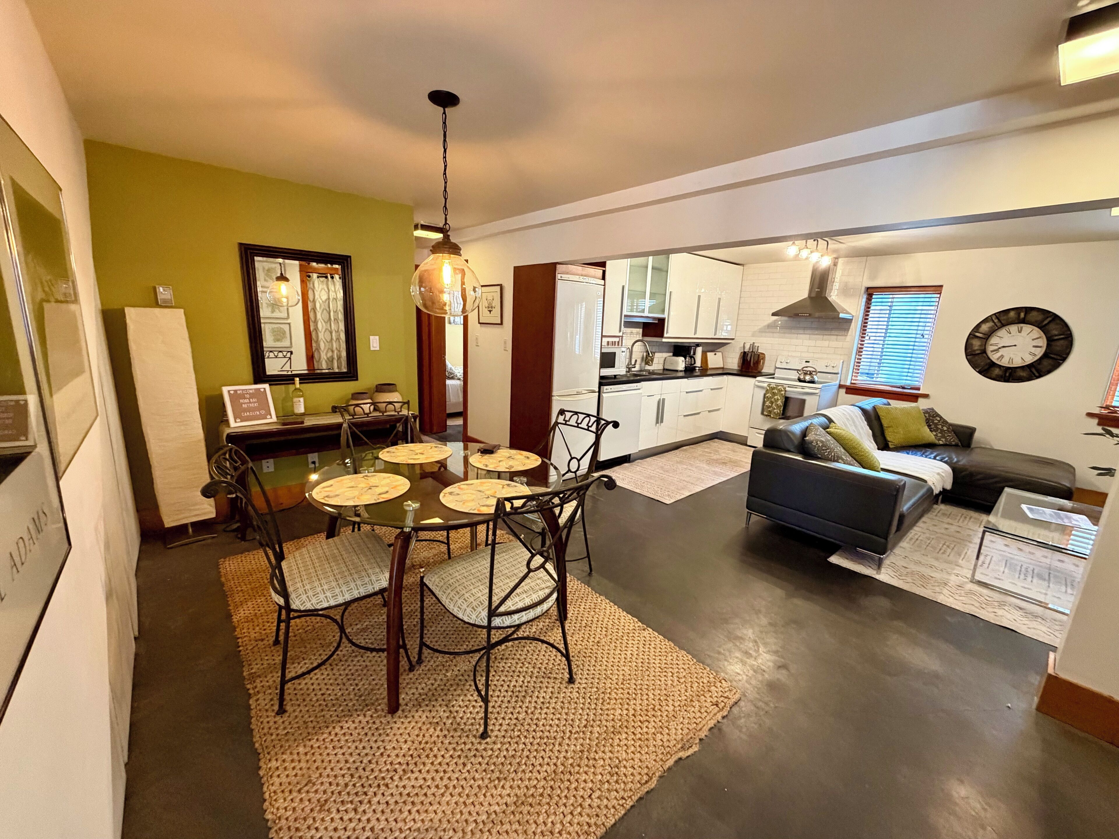 Eine moderne Küche mit weißen Schränken, ein Essbereich mit Glastisch und Stühlen sowie ein Wohnzimmer mit Sofa und Wanduhr.