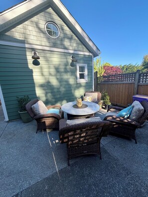 Une terrasse dotée de meubles en osier et d’une table ronde, une petite fenêtre et un luminaire sur un bâtiment vert.