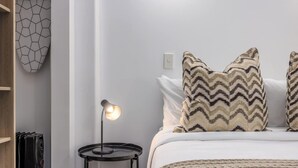 Una cama bien hecha con almohadones estampados, una lámpara de noche y un interruptor de luz en la pared.
