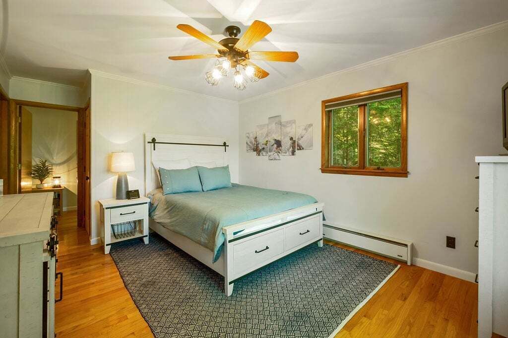 A bedroom with a bed, nightstand, lamp, ceiling fan, window, and artwork on the wall.