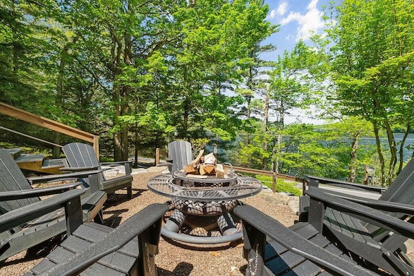 A deck with outdoor seating and a fire pit, surrounded by trees and overlooking a body of water. - Spacious 7BR retreat with hot tub, fire pit, dock, and year-round views (Swanton)