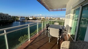 Un balcon avec une table et des chaises, une rambarde en verre et une vue sur une étendue d’eau et des bâtiments.
