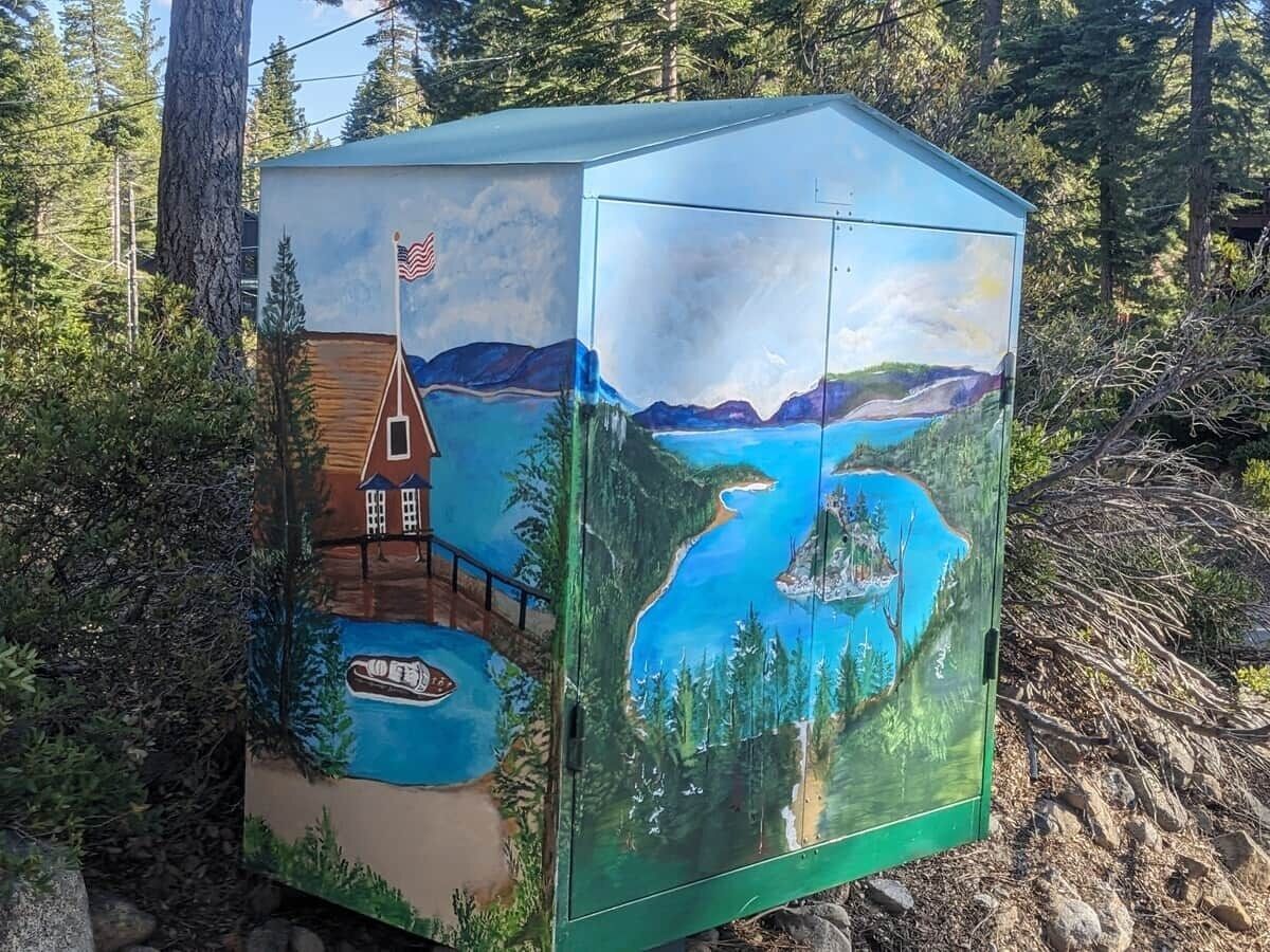 A green storage box with a painting of a cabin by a lake surrounded by trees.