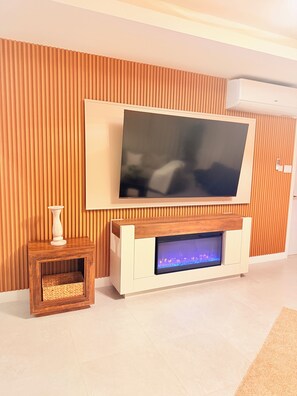 A modern living room with a flat-screen TV mounted on a wooden wall, a fireplace with a glass front, and a small wooden side table with a vase. - Spacious 2 bedroom apartment in charming Kingston with pool. (Kingston)