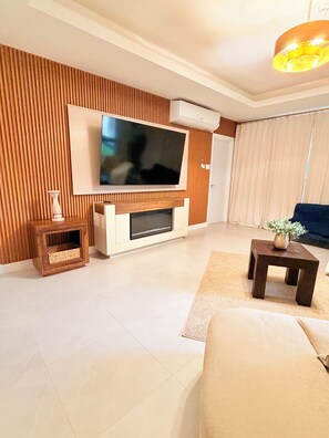 A modern living room with a flat-screen TV mounted on a wooden wall, a sofa, a coffee table with a plant, and a small side table. - Spacious 2 bedroom apartment in charming Kingston with pool. (Kingston)
