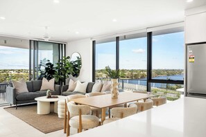Ein modernes Wohnzimmer mit Sofa, Couchtisch und Essbereich, große Fenster mit Blick auf ein Gewässer sowie eine Küche mit Kühlschrank.