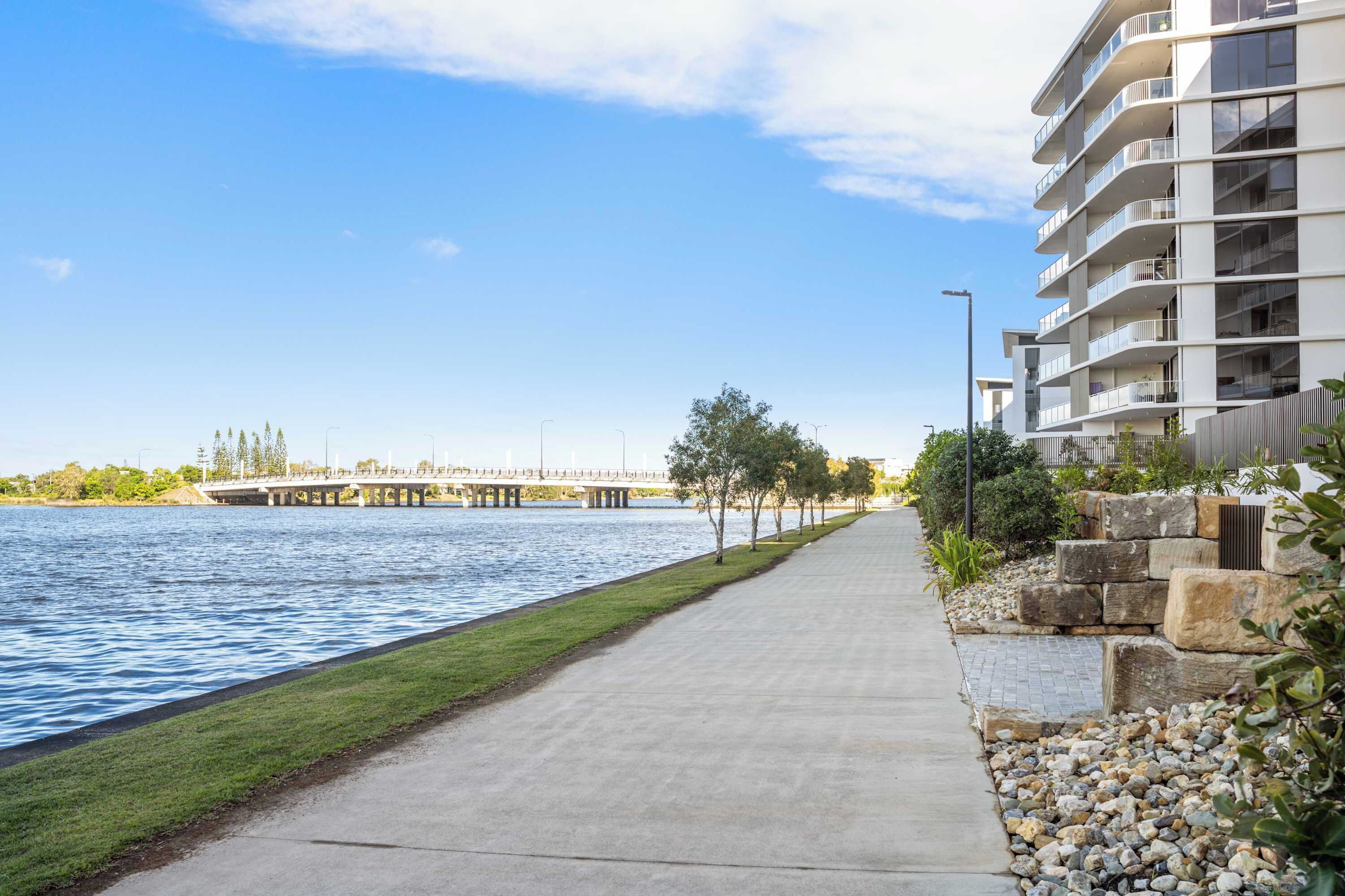 A waterfront walkway with a bridge in the background, a modern building with balconies, and a grassy area with rocks and plants.