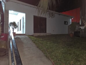 Ein Gebäude mit Außenansicht bei Nacht, ein Betonweg führt zu einer offenen Tür, ein Blumentopf und ein roter Zaun sind zu sehen.