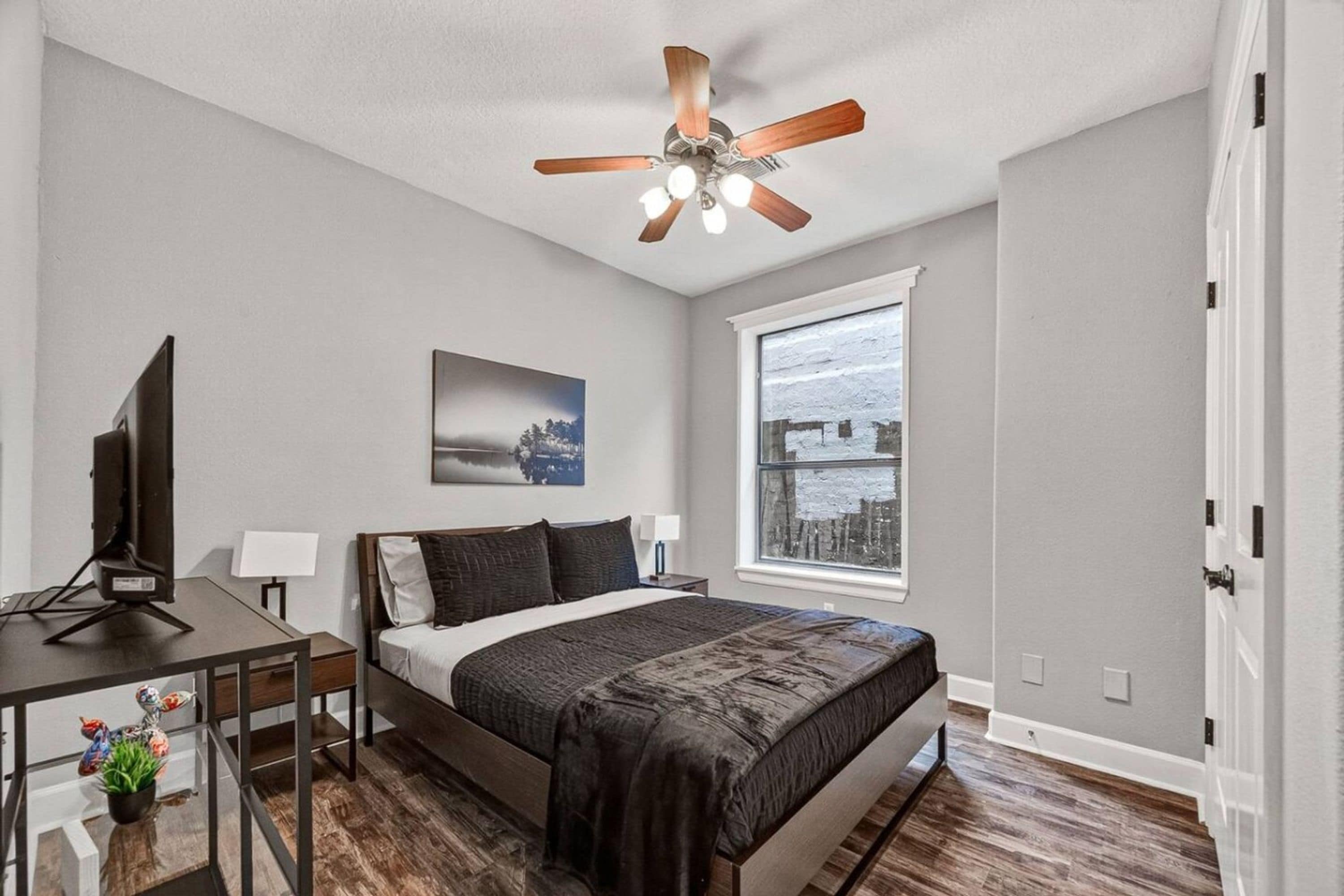 A bedroom with a bed, a desk, a television, a ceiling fan, a window with a view, and a painting on the wall.