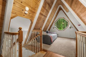 Ein Schlafzimmer mit schrägen Wänden, einer Holztreppe, einem Bett und einem Fenster mit Blick auf Grünflächen.