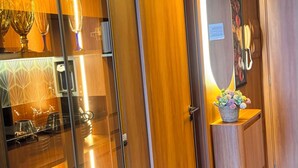 Habitación de hotel con un armario de madera, una vitrina de cristal con copas de vino, un espejo y una pequeña mesa con un arreglo floral.