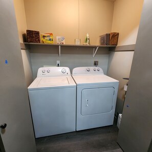 Ein Waschraum mit zwei Waschmaschinen mit Frontlader, einem Regal für Waschmittel und einem kleinen Bügelbrett.