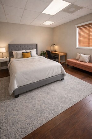 A bedroom with a bed, a desk, a chair, a bench, a lamp, and a window with blinds.