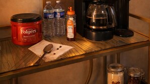 Preparación de una barra de café con una cafetera Folgers, botellas de agua, un vaso de jugo y una cuchara sobre una mesa de vidrio.