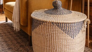 Una cesta redonda tejida con tapa, sobre un suelo de madera en una habitación con un sofá y una lámpara.