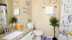 Ein Badezimmer mit Waschbecken, Toilette und Duschvorhang. An den Wänden hängen Bilder in Rahmen und ein dekoratives Schild ist angebracht.