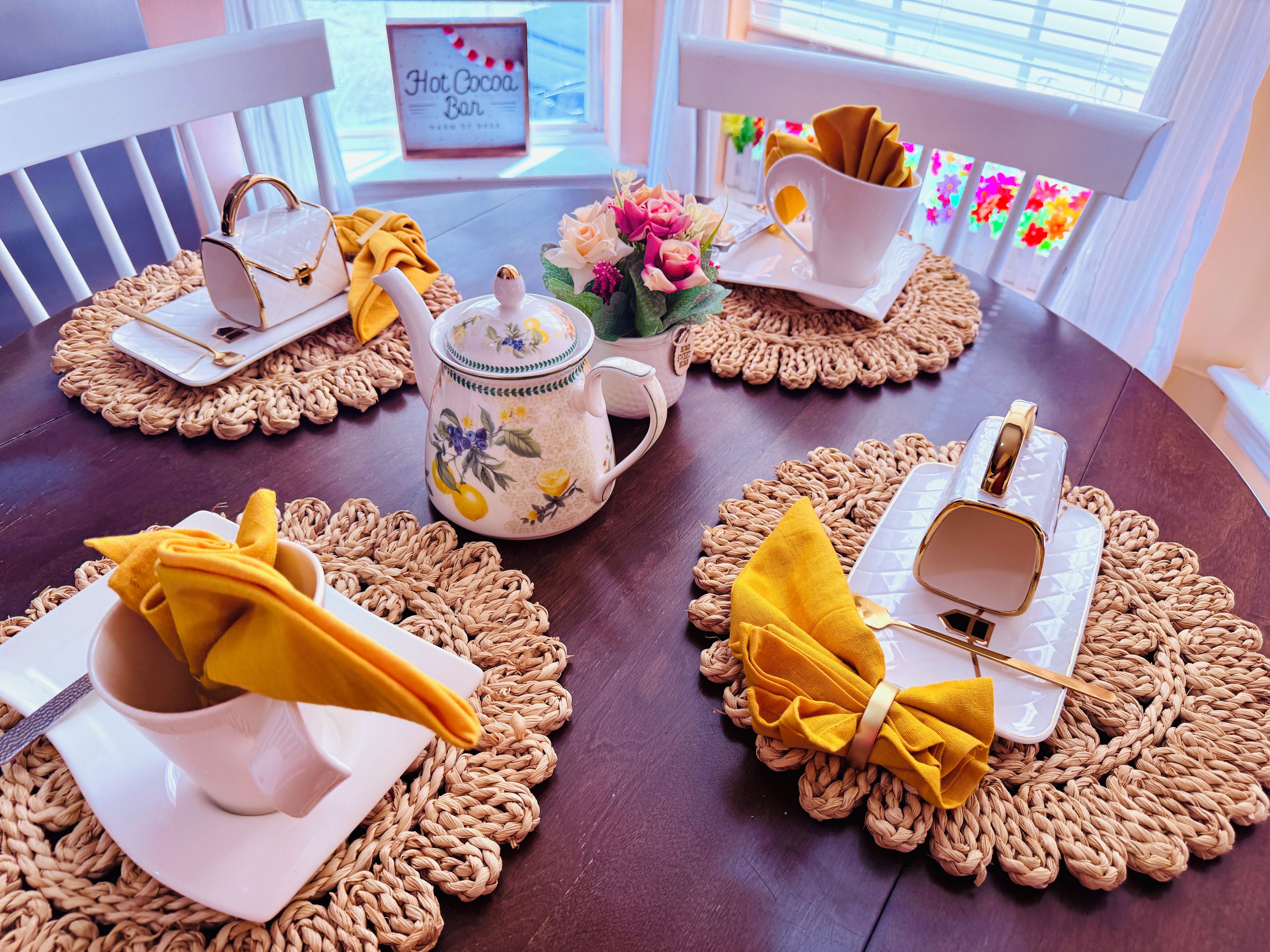 Una mesa de comedor dispuesta con individuales, una tetera, tazas y un letrero que dice 'Hot Cocoa Bar'.
