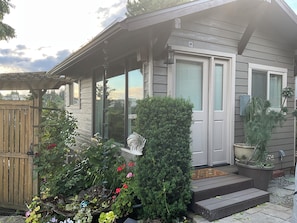 Ein Haus mit überdachter Veranda, einem kleinen Garten mit verschiedenen Pflanzen und Blumen sowie einem sichtbaren Fenster.