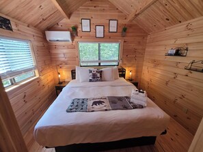 Un dormitorio de cabaña de madera acogedor, con una cama, dos mesitas de noche, un ventanal con persianas y cuadros enmarcados en la pared.