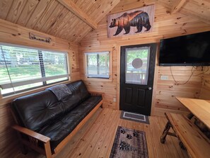 Interior de una cabaña con un sofá de cuero negro, un colgador temático de osos, un televisor montado en la pared y piso de madera.