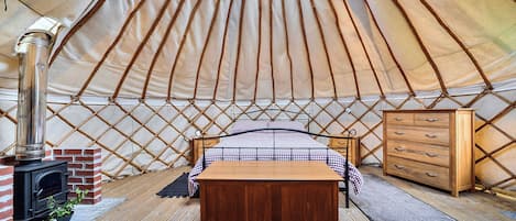 A cozy interior of a yurt with a bed, a dresser, and a wood-burning stove.