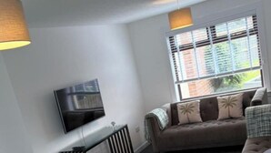 A living room with a sofa, a television mounted on the wall, a dining table with chairs, and a window with blinds. - Ayr 2-Bed House, Sleeps 4 with Garden & Parking (Ayr)