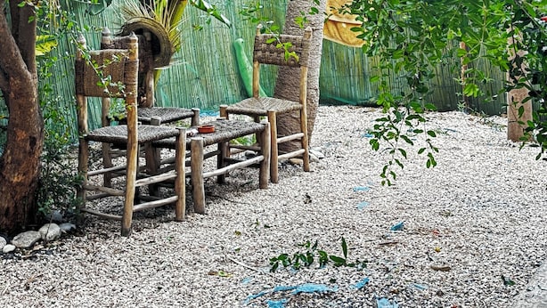 A rustic outdoor seating area with wooden chairs and a woven table, surrounded by lush greenery and a bamboo fence.