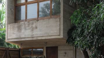 A two-story concrete building with large windows, a wooden door, and a small garden area with plants.